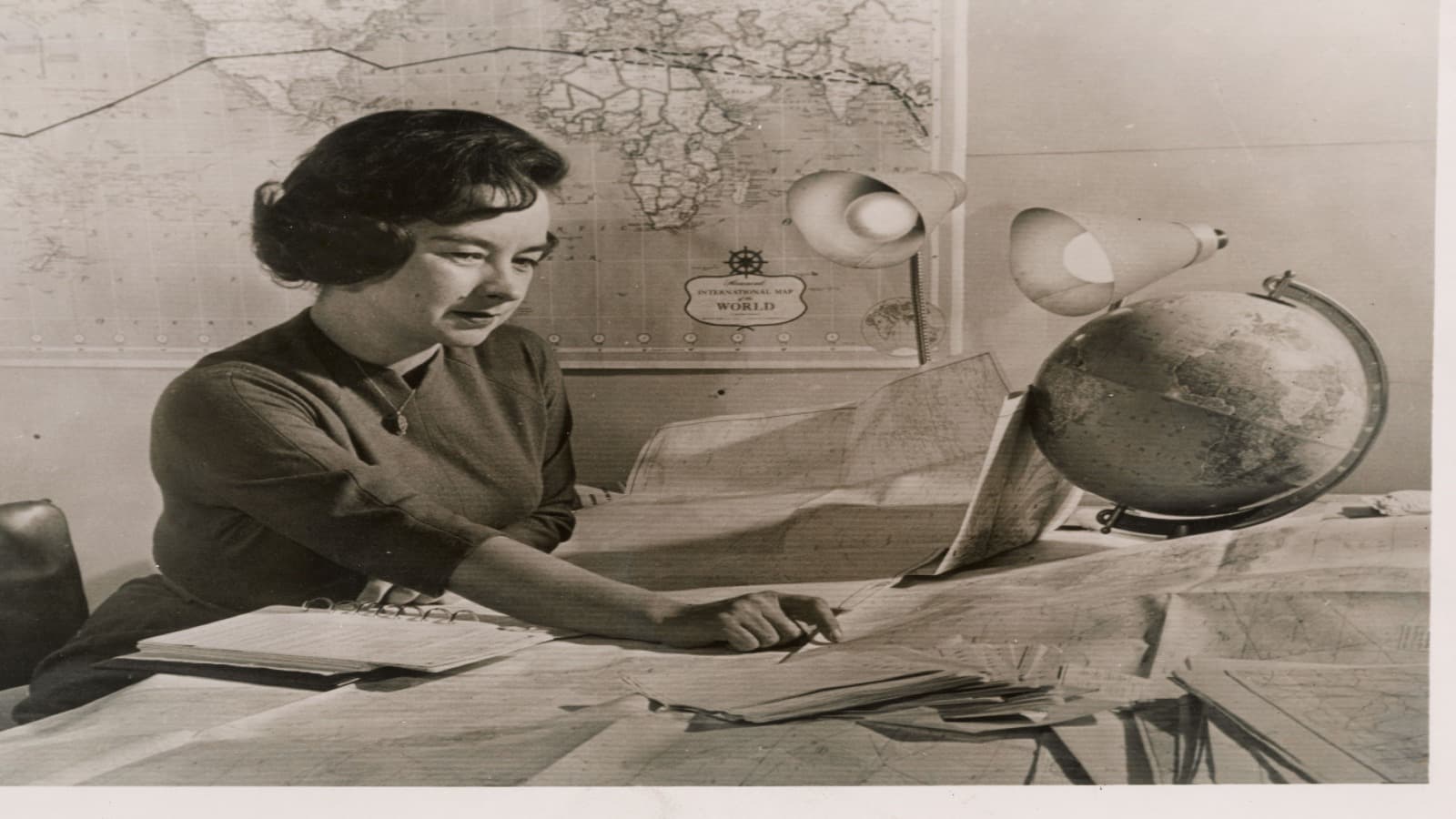 Jerrie Mock étudie des cartes à côté d'un globe terrestre et d'une carte du monde alors qu'elle prépare son tour du monde en solitaire, janvier 1964.