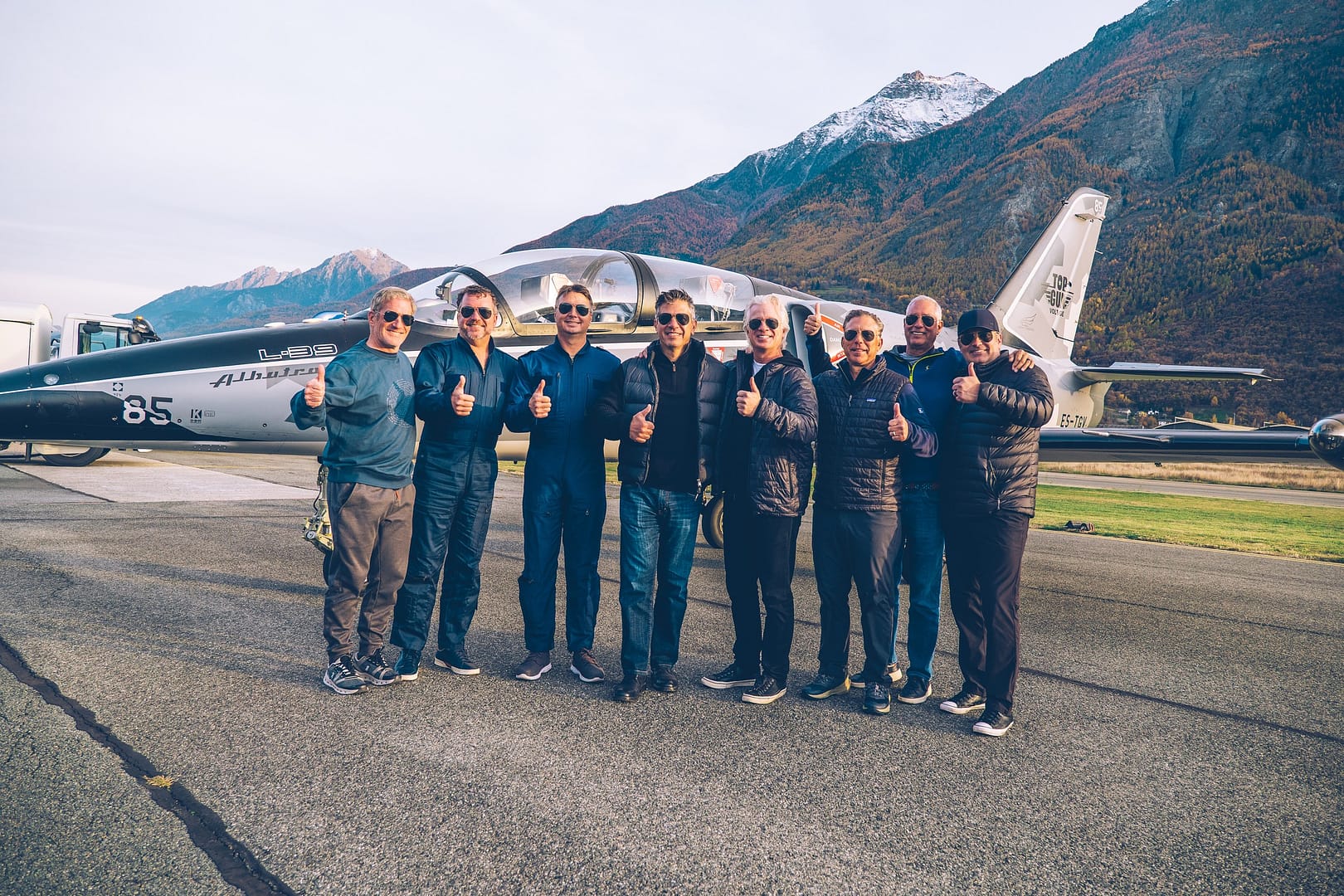 A group of friends in flight suits give a thumbs-up in front of a MiGFlug L-39 Albatros jet, with the Alps behind them at Aosta