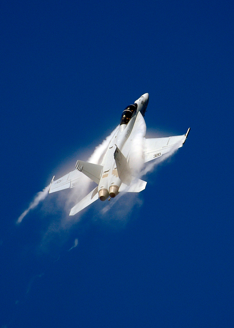 An F/A-18F Super Hornet making a high-speed pass, vapour tearing off the wings.