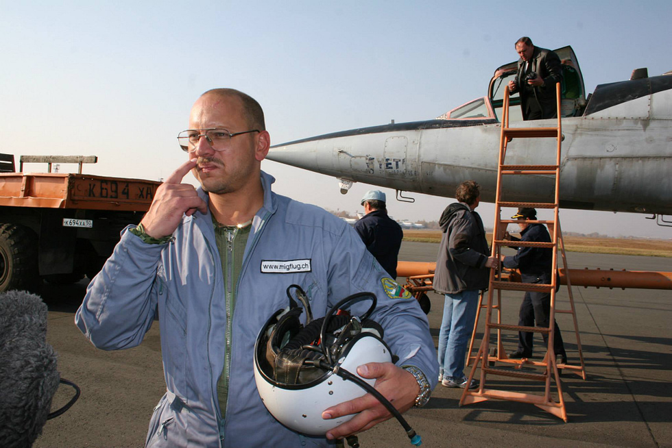 A MiGFlug pilot in flight suit holding a helmet beside a MiG-25